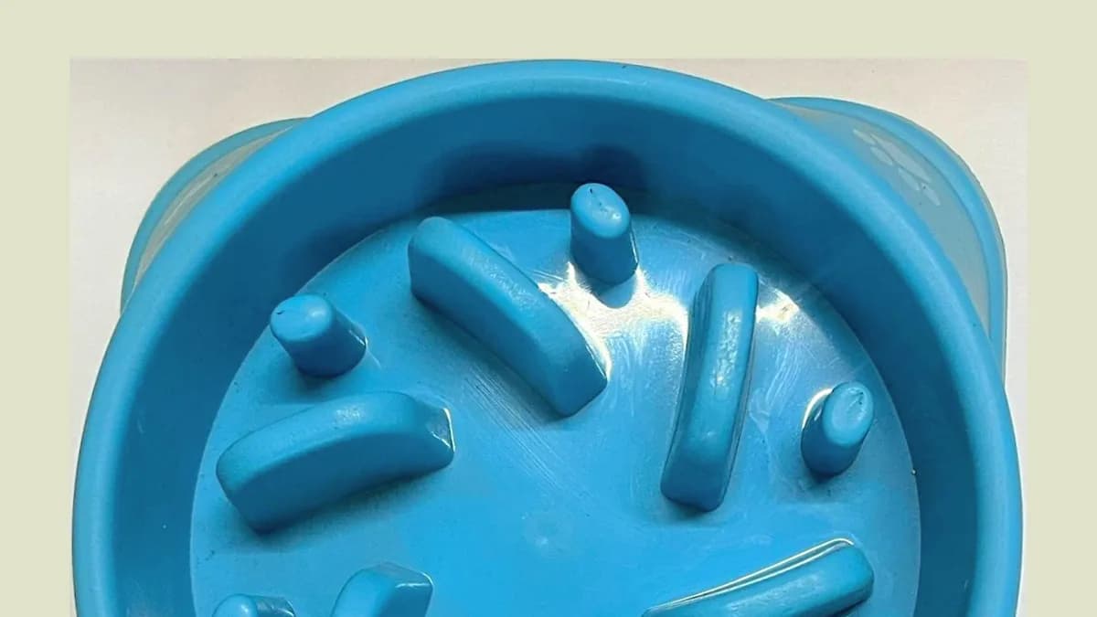 A top-down view of a square, light blue slow feeder dog bowl. The interior features a challenging arrangement of raised studs and curved ridges that create separate compartments and pathways. This com